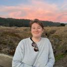 Jocelyn, a young white person with short hair, smiles in front of a pink sunset